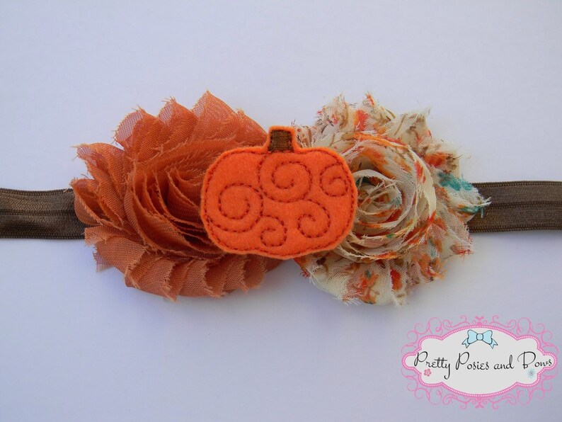 May include: A brown and cream fabric headband with a felt orange pumpkin applique. The pumpkin has a swirl design.