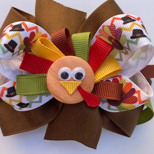 May include: A brown, green, red, and yellow turkey-themed hair bow with a wooden turkey head in the center. The turkey head has googly eyes and a yellow beak.