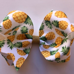 May include: A white hair bow with a pineapple print. The bow is made of a soft, textured fabric and has a gold center.