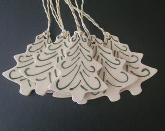 May include: A collection of ceramic Christmas tree ornaments. Each ornament is off-white with a green tree design and a gold and white string for hanging. The ornaments are arranged on a dark surface.