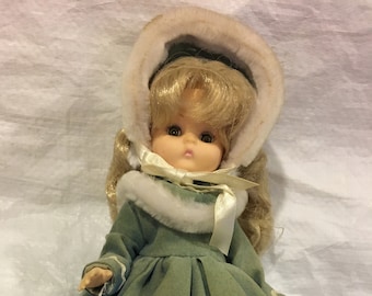 May include: A vintage doll wearing a green velvet dress with a white fur trim and a white fur hat. The doll has blonde hair and brown eyes. The doll is holding a tag that says "Currier and Ives" and "Effanbee Doll Company".