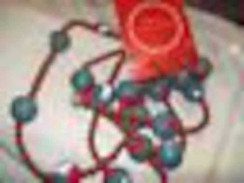 May include: A necklace with teal beads on a reddish-brown cord. The beads are spaced along the cord, creating a distinctive pattern. A red tag with a circular design is visible in the background.