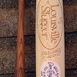 May include: A wooden Louisville Slugger baseball bat with a white label that reads "Genuine Louisville Slugger" and "Hall of Fame Don Drysdale Golf Classic 1989". The bat is 125 cm long and has a brown wooden handle.