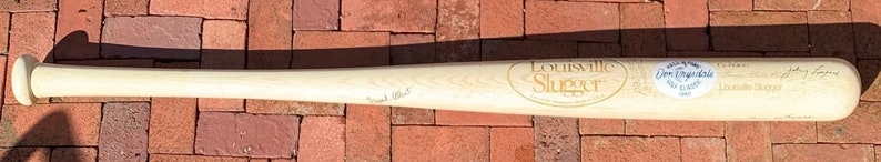 May include: A wooden Louisville Slugger baseball bat with signatures from various players, including "Johnny Bench" and "Charlie Hustle". The bat is signed with a black marker and has a white sticker with the text "Don Drysdale 1962" on the side.