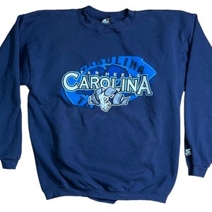 May include: A navy blue sweatshirt with a large graphic of the University of North Carolina Tar Heels logo. The logo features the words "Tar Heels" and "Carolina" in a blue and white font, with a blue mascot in the center.