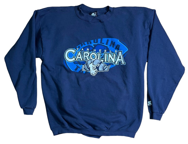 May include: A navy blue sweatshirt with a large graphic of the University of North Carolina Tar Heels logo. The logo features the words "Tar Heels" and "Carolina" in a blue and white font, with a blue mascot in the center.