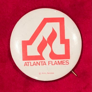 May include: A white button with a red Atlanta Flames logo. The logo features a stylized "A" with flames inside. The text "Atlanta Flames" is below the logo. The text "NHL Services" is in smaller font below the text "Atlanta Flames".