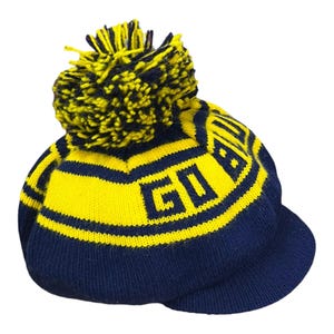 May include: A navy blue and yellow knit beanie with a pom-pom on top and a short brim. The hat features horizontal stripes and the text GO BLUE knitted into the fabric in a bold, block font. The pom-pom is a mix of navy blue and yellow yarn.
