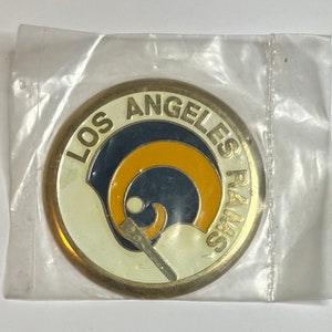 May include: A gold-colored metal coin with a white background and the words "Los Angeles Rams" in a circle around a blue and gold ram's head logo.
