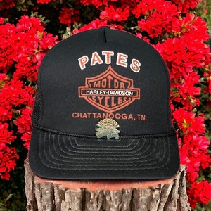 May include: Black trucker hat with a white "PATES" logo and a Harley-Davidson emblem. The hat also has a gold "HOG" pin with a Harley-Davidson logo and the text "Chattanooga, TN".