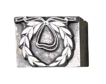 May include: A silver-colored metal stamp with a pear-shaped design surrounded by a laurel wreath and a ribbon at the top. The stamp is rectangular and has a textured surface.