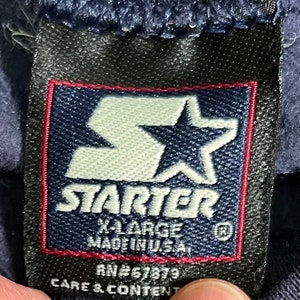 May include: A navy blue Starter brand tag with a white star logo and the text "Starter" in white. The tag also includes the text "X-Large Made in U.S.A." and "RN#67879 Care & Content".