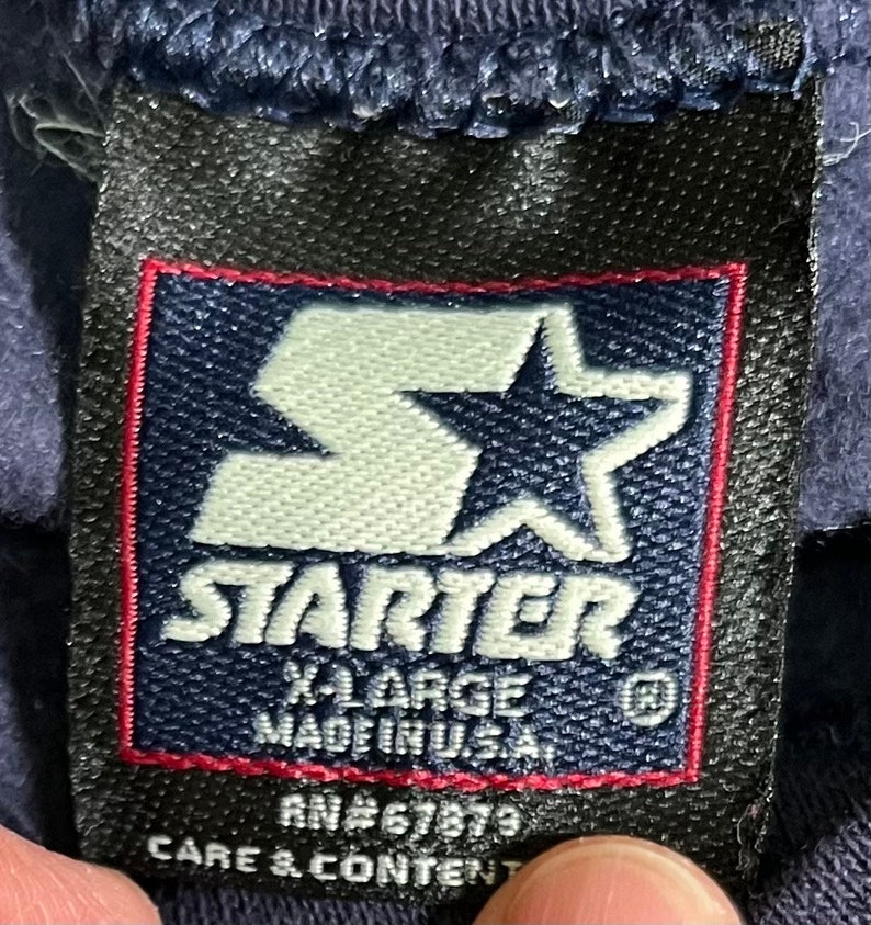 May include: A navy blue Starter brand tag with a white star logo and the text "Starter" in white. The tag also includes the text "X-Large Made in U.S.A." and "RN#67879 Care & Content".