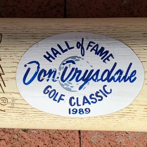 May include: A white oval sticker with blue text that reads "Hall of Fame Don Drysdale Golf Classic 1989". The sticker is on a wooden baseball bat with the text "Donruss" and "Genuine Story Louis" engraved on the wood.