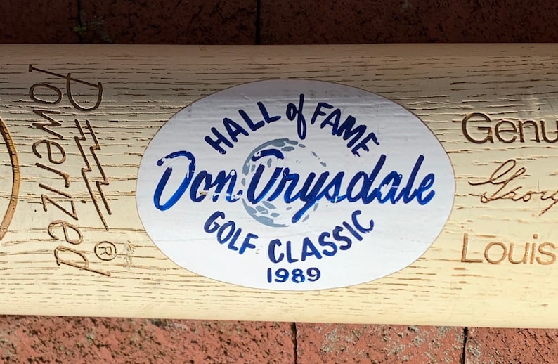 May include: A white oval sticker with blue text that reads "Hall of Fame Don Drysdale Golf Classic 1989". The sticker is on a wooden baseball bat with the text "Donruss" and "Genuine Story Louis" engraved on the wood.