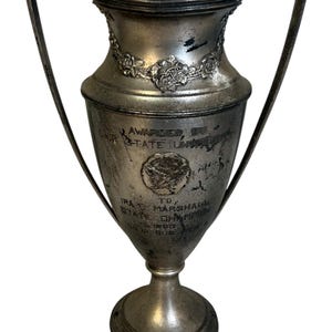 May include: A vintage silver trophy with two handles, featuring a floral garland around the top. The trophy has inscriptions, including "Awarded by OUR STATE UNIVERSITY" and "IRA T. MARSHALL STATE CHAMPION". The base is round and the trophy is likely from 1953.