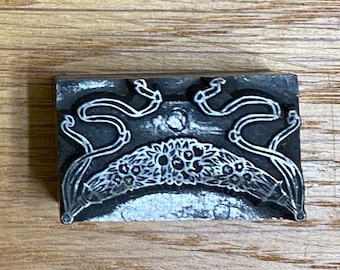 May include: A rectangular, dark gray printing block with a floral and ribbon design. The design features a curved floral arrangement at the bottom and flowing ribbons at the top. The block is made of metal and sits on a wooden surface.