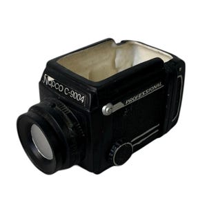 May include: Black ceramic ashtray shaped like a vintage camera. The ashtray has a rectangular shape with a lens on the front and the text "Napco C-9004" and "PROFESSIONAL" printed on the side. The interior is beige.