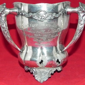 May include: A silver trophy cup with two ornate handles. The cup has an inscription that reads "Alfred Pero Jr. Orchard and Basket Ball Meet 1903 - 1904 For Highest Percentage in Basket Throwing Contest"