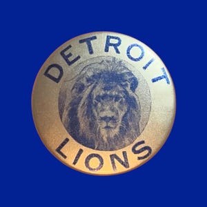 May include: A round, gold-colored button with the words "DETROIT LIONS" in blue lettering around the edge. The center features a detailed illustration of a lion's head. The button is set against a solid blue background.