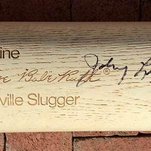 May include: A white wooden Louisville Slugger baseball bat with the text "Genuine George "Babe" Ruth" and "Louisville Slugger" engraved on the surface. The bat is signed by "Johnny Bench".