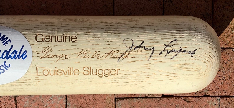 May include: A white wooden Louisville Slugger baseball bat with the text "Genuine George "Babe" Ruth" and "Louisville Slugger" engraved on the surface. The bat is signed by "Johnny Bench".