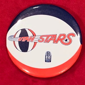 May include: A white, red, and blue button with a basketball and the text "UTAH STARS" in red with a star. The button also has a small logo that says "AMERICAN BASKETBALL ASSOCIATION".