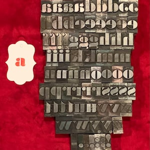 May include: A collection of vintage metal letterpress printing blocks in various sizes and styles. The blocks are arranged in a vertical stack, with the letter 'a' at the top and the letter 'o' at the bottom. The blocks are made of a dark metal and have a worn, aged appearance.