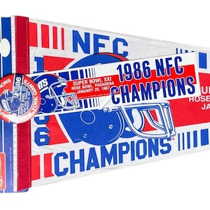 May include: A vintage New York Giants pennant and button set. The pennant is white with blue and red accents, featuring the text "1986 NFC CHAMPIONS" and a football helmet graphic. The button also displays the team logo and championship details.