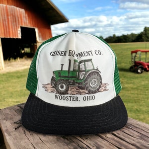 May include: A white and green trucker hat with a black bill. The front of the hat features a green tractor with the text "Geiser Equipment Co. Wooster, Ohio".