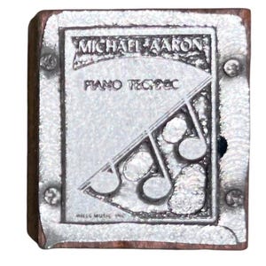 May include: A rectangular, silver-colored metal stamp with the text "MICHAEL AARON PIANO TECHNIQUE" and a musical note design. The stamp is mounted on a wooden block and secured with small rivets. The words "MILLS MUSIC INC" are also visible.