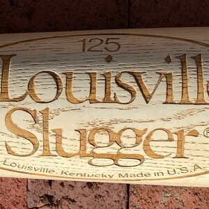 May include: A white wooden baseball bat with a brown oval logo that says "Louisville Slugger" with the text "Louisville, Kentucky Made in U.S.A." below the logo. The number "125" is above the logo.