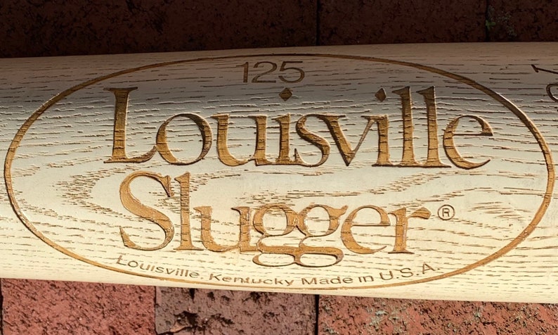May include: A white wooden baseball bat with a brown oval logo that says "Louisville Slugger" with the text "Louisville, Kentucky Made in U.S.A." below the logo. The number "125" is above the logo.