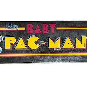 May include: A vintage Baby Pac-Man arcade game marquee. The black background features the words "Bally BABY PAC-MAN" in yellow and red lettering. Cartoon Pac-Man characters are on either side of the text, with a yellow Pac-Man character on the left and a blue ghost on the right.