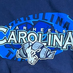 May include: A navy blue t-shirt with a blue and white graphic of the University of North Carolina Tar Heels logo. The logo features the words "Tar Heels" and "Carolina" with a ram mascot.
