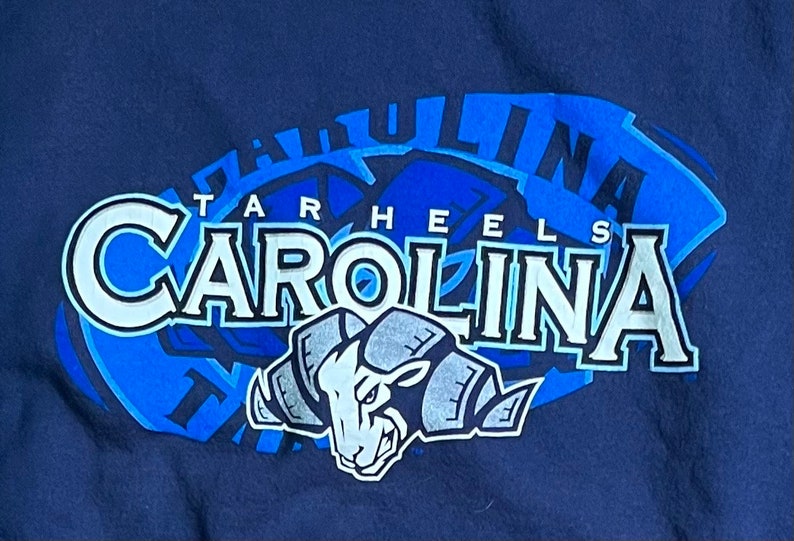May include: A navy blue t-shirt with a blue and white graphic of the University of North Carolina Tar Heels logo. The logo features the words "Tar Heels" and "Carolina" with a ram mascot.