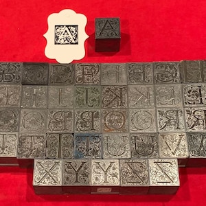 May include: A set of 26 metal letterpress printing blocks, each featuring a different letter of the alphabet in a decorative, ornate font. The letters are arranged in alphabetical order and are all the same size. The blocks are made of a dark gray metal and have a slightly textured surface.
