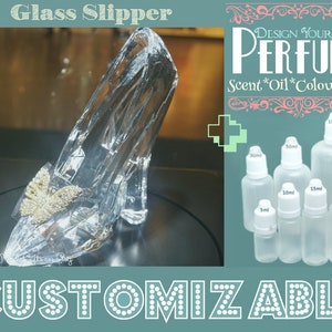 Puede incluir: Una zapatilla de cristal con un diseño de mariposa. La imagen también muestra un conjunto de botellas de vidrio transparente de diferentes tamaños, etiquetadas con medidas en mililitros. El texto "DESIGN YOUR OWN PERFUME" se muestra encima de las botellas. El texto "Scent*Oil*Colour*Size" se muestra debajo de las botellas. El texto "CUSTOMIZABLE" se muestra en la parte inferior de la imagen.