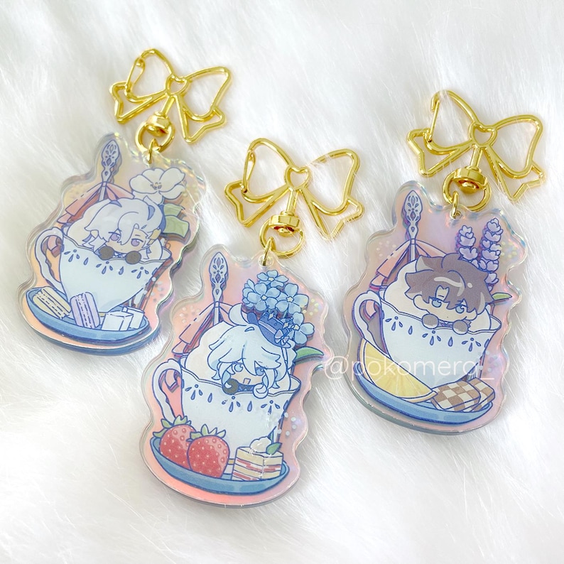 May include: Three iridescent keychains featuring cartoon characters in teacups. Each keychain has a gold bow and a gold chain. The characters are a white haired person with blue flowers, a white haired person with strawberries, and a brown haired person with lavender.