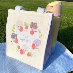 May include: A cream-colored lunch bag with a cartoon design of cat-like characters holding strawberries, surrounded by a floral wreath. The text "bon appétit" is in the center. A yellow water bottle is next to the bag.