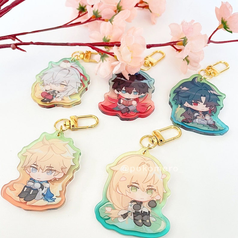 Pode incluir: Cinco chaveiros de personagens de anime coloridos com correntes douradas. Cada chaveiro apresenta um personagem diferente com um design e esquema de cores &uacute;nicos. Os chaveiros s&atilde;o exibidos em um fundo branco com flores de cerejeira rosa.