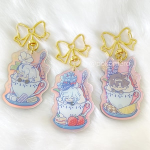May include: Three iridescent keychains featuring cartoon characters in teacups. Each keychain has a gold bow and a gold chain. The characters are surrounded by flowers and sweets.