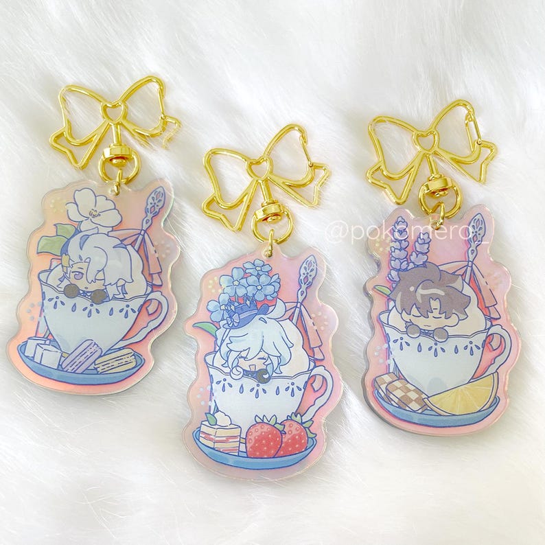 May include: Three iridescent keychains featuring cartoon characters in teacups. Each keychain has a gold bow and a gold chain. The characters are surrounded by flowers and sweets.