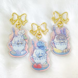 May include: Three gold charm keychains with colorful character illustrations. Each keychain features a character in a teacup with a dessert on a plate. The characters are a blonde woman with a blue flower, a brunette man with a purple flower, and a blonde woman with a blue flower.