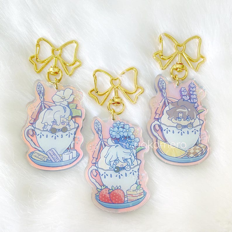 May include: Three gold charm keychains with colorful character illustrations. Each keychain features a character in a teacup with a dessert on a plate. The characters are a blonde woman with a blue flower, a brunette man with a purple flower, and a blonde woman with a blue flower.