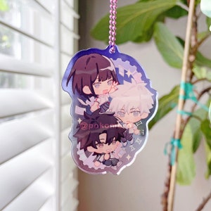 May include: A clear acrylic keychain featuring three anime characters. The characters are a girl with long black hair, a boy with white hair, and a boy with black hair. The keychain is decorated with glitter and stars. The text "@pokomi_o" is visible on the keychain.