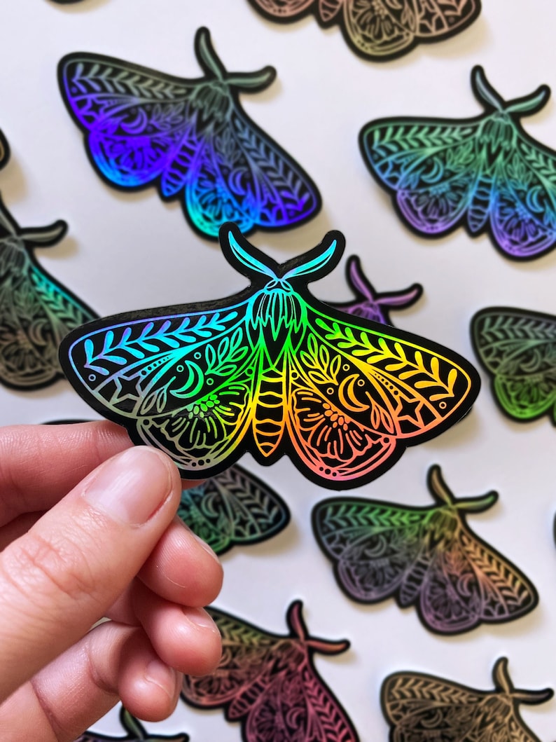 May include: A set of iridescent holographic stickers featuring a detailed moth design with intricate floral patterns and crescent moons. The stickers are black with a rainbow sheen.