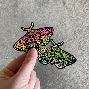 May include: Two iridescent holographic stickers of moths with intricate black and white designs. The moths have a rainbow gradient colour scheme.