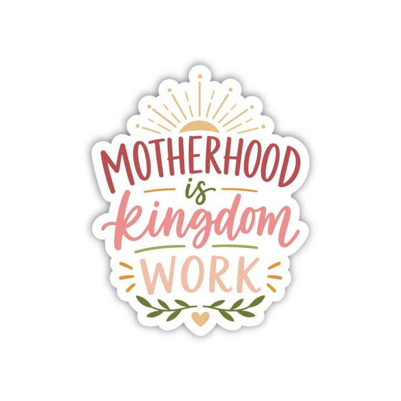 以下が含まれることがあります： ピンク、赤、緑のスクリプトで「MOTHERHOOD is kingdom WORK」という言葉が書かれた白いステッカー。デザインには、サンバースト、葉、ハートが含まれています。