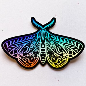May include: A rainbow iridescent sticker of a moth with intricate floral and moon designs. The moth is black with white details and a rainbow gradient.